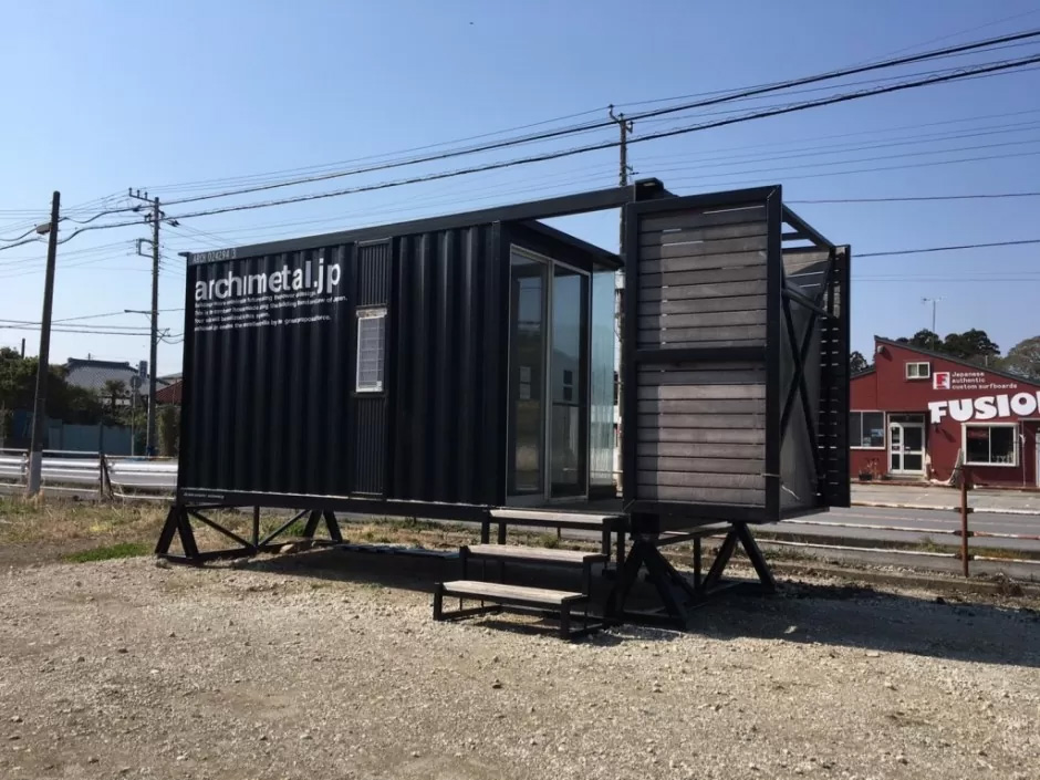 006 - あなたがデザインする新しい建築のかたち Modular container building on stilts with steps, featuring archmetal.jp signage, beside a road.