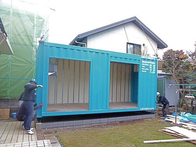 Two construction workers are installing a blue shipping-container module on a raised foundation beside a house under construction.