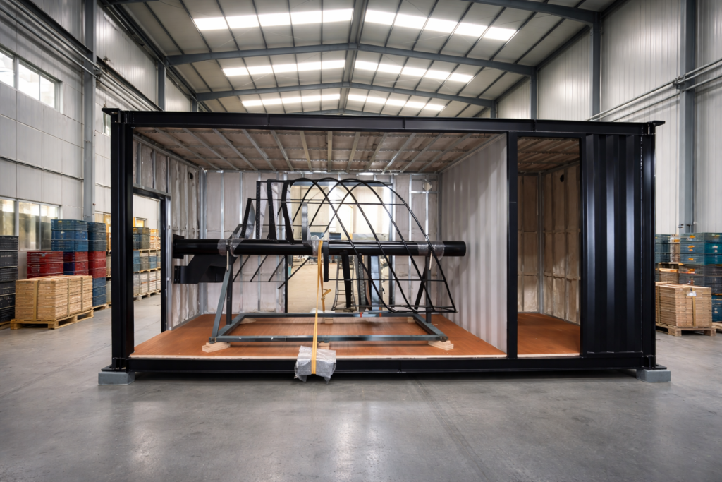 Industrial shipping container opened in a warehouse, showing a large black metal frame and mechanical rig mounted on a wooden platform inside the container.