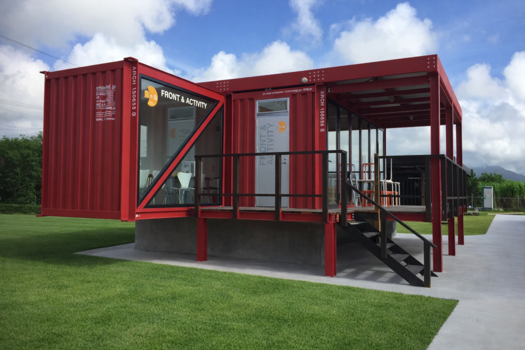 Red modular building with glass walls and a raised deck labeled Front & Activity, set on a grassy field.