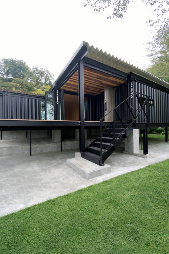 Modern black metal building on stilts with a metal staircase leading to an elevated deck and a corrugated roof, set on a concrete base with green lawn nearby.
