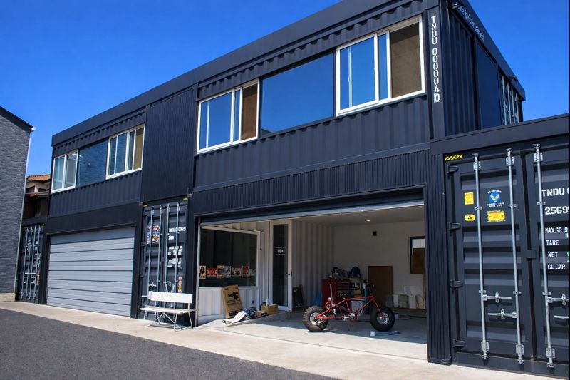 Two-story black shipping-container building with an open ground-floor garage showing a red bike and scattered items on a sunny day.