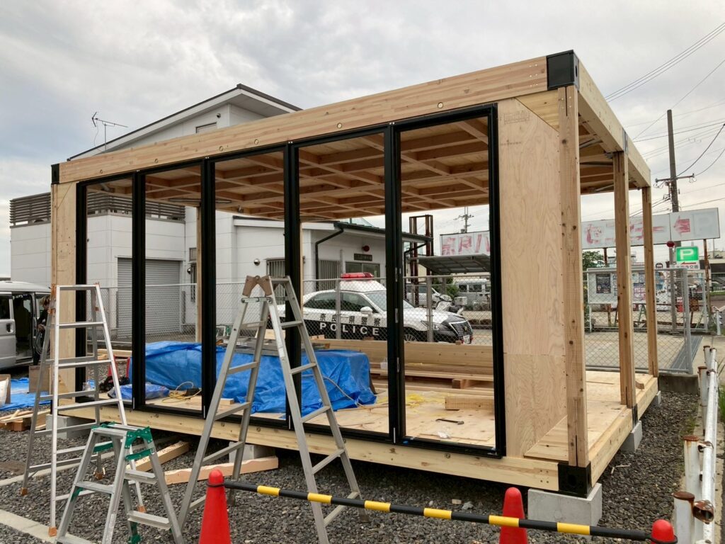 D6745C5E-DEFA-4DEF-8ADB-3C0A94DCD304 - あなたがデザインする新しい建築のかたち Wood-framed outdoor structure under construction with large black-framed glass panels and a wooden deck, tools and ladders nearby.