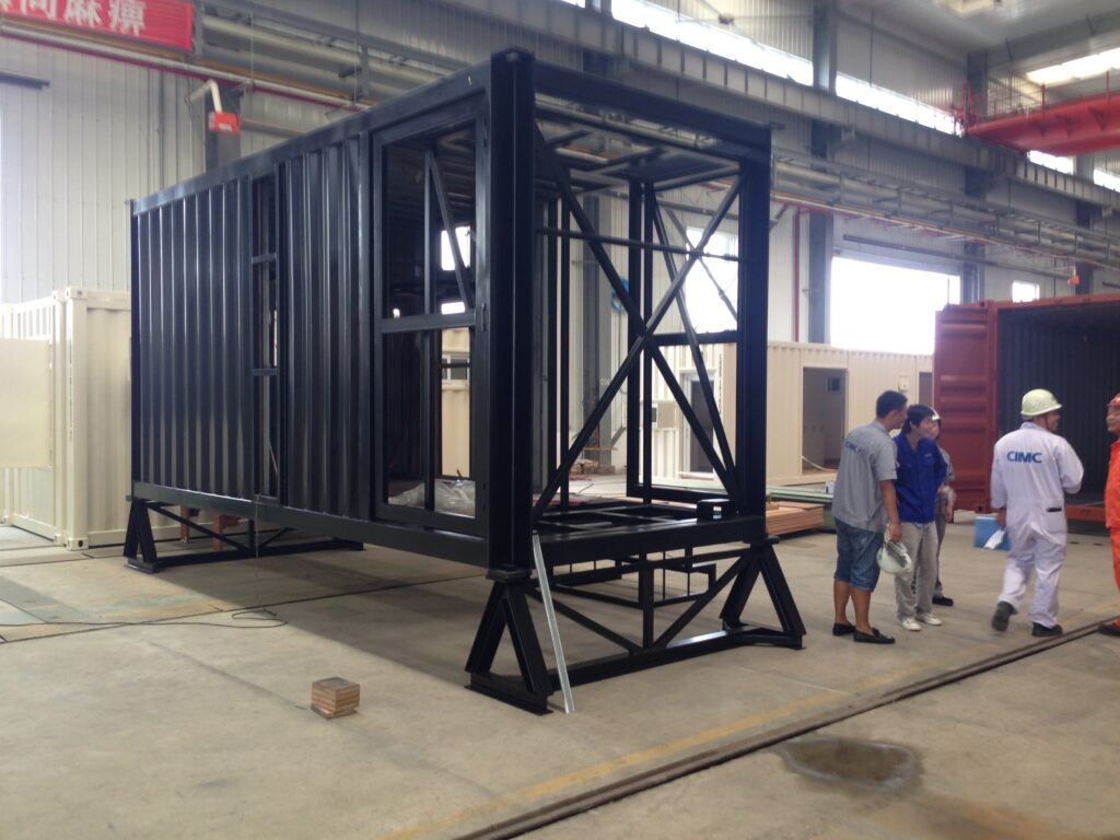 Large black steel frame structure being assembled in an industrial workshop, with workers nearby and a red overhead crane above.