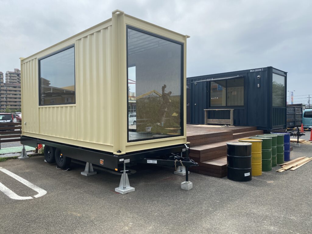 Beige modular container with large glass window sits on a trailer in a parking lot, with a dark blue container and wooden decking nearby.