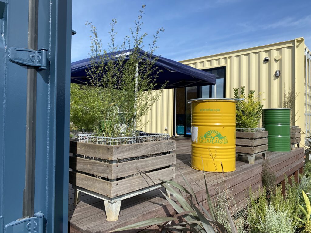 Outdoor industrial area with wooden planter boxes, tall plants, and a navy canopy in front of a beige shipping-container building.