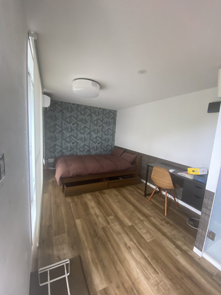 IMG_7792 - あなたがデザインする新しい建築のかたち Modern bedroom with brown wooden bed, geometric wallpaper accent wall, and a desk with chair by the wall.