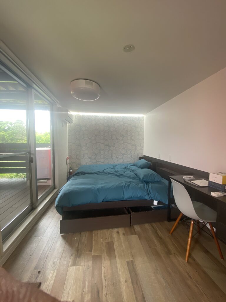 IMG_7800 - あなたがデザインする新しい建築のかたち Modern bedroom with a low platform bed in blue linens, a desk and chair, and a sliding glass door to a balcony on the left. The wall behind the bed has a circular pattern design and wood flooring runs throughout.
