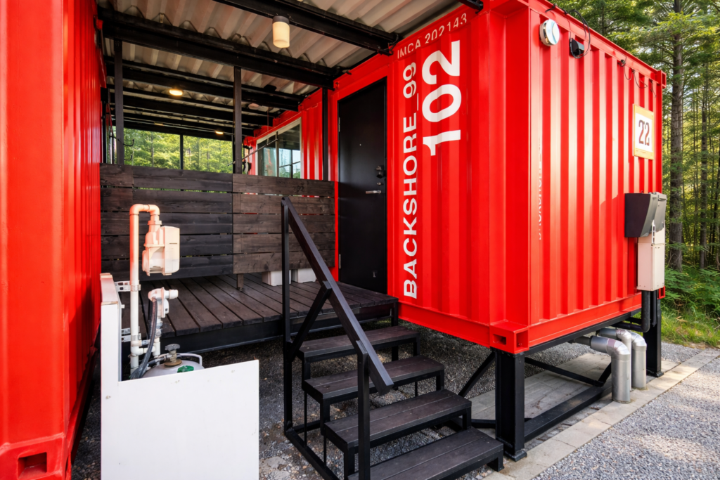 d243156a-b4ad-4549-a202-5578a08f038b - あなたがデザインする新しい建築のかたち Red shipping-container building with a dark wooden deck, stairs, and a black door, surrounded by trees in a forest setting.