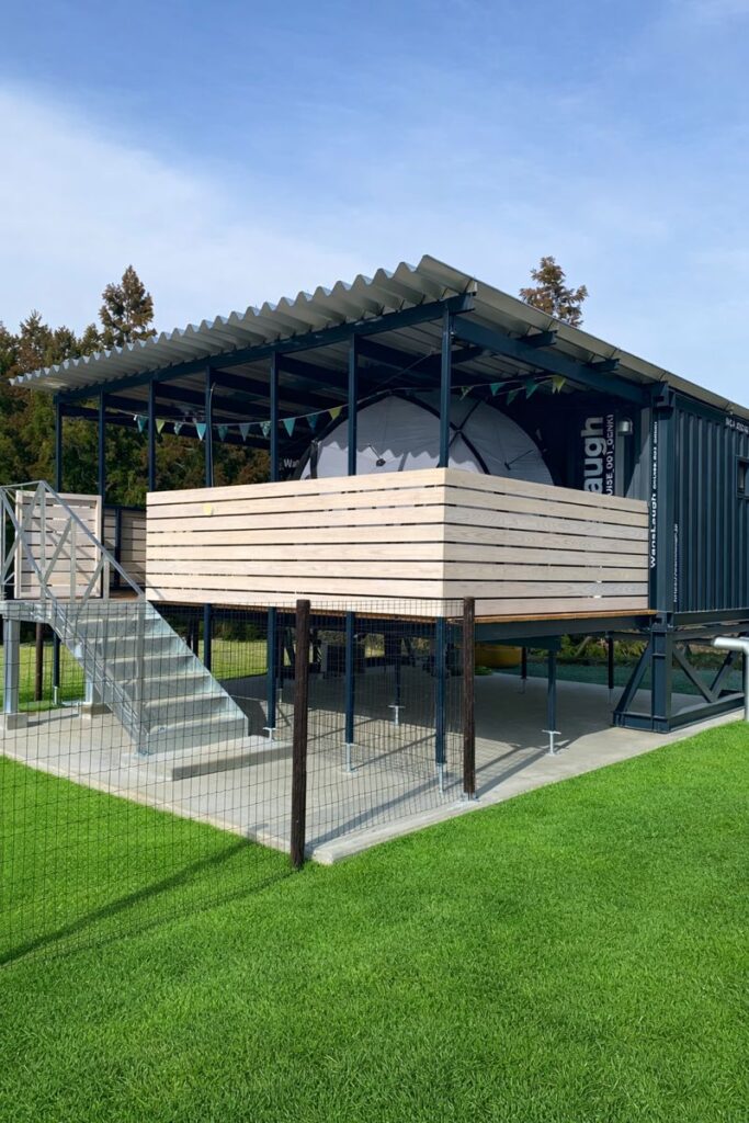 Blue shipping-container shelter on stilts with a wooden deck, metal stairs, and a fenced grassy area; a white tent is inside the deck enclosure.