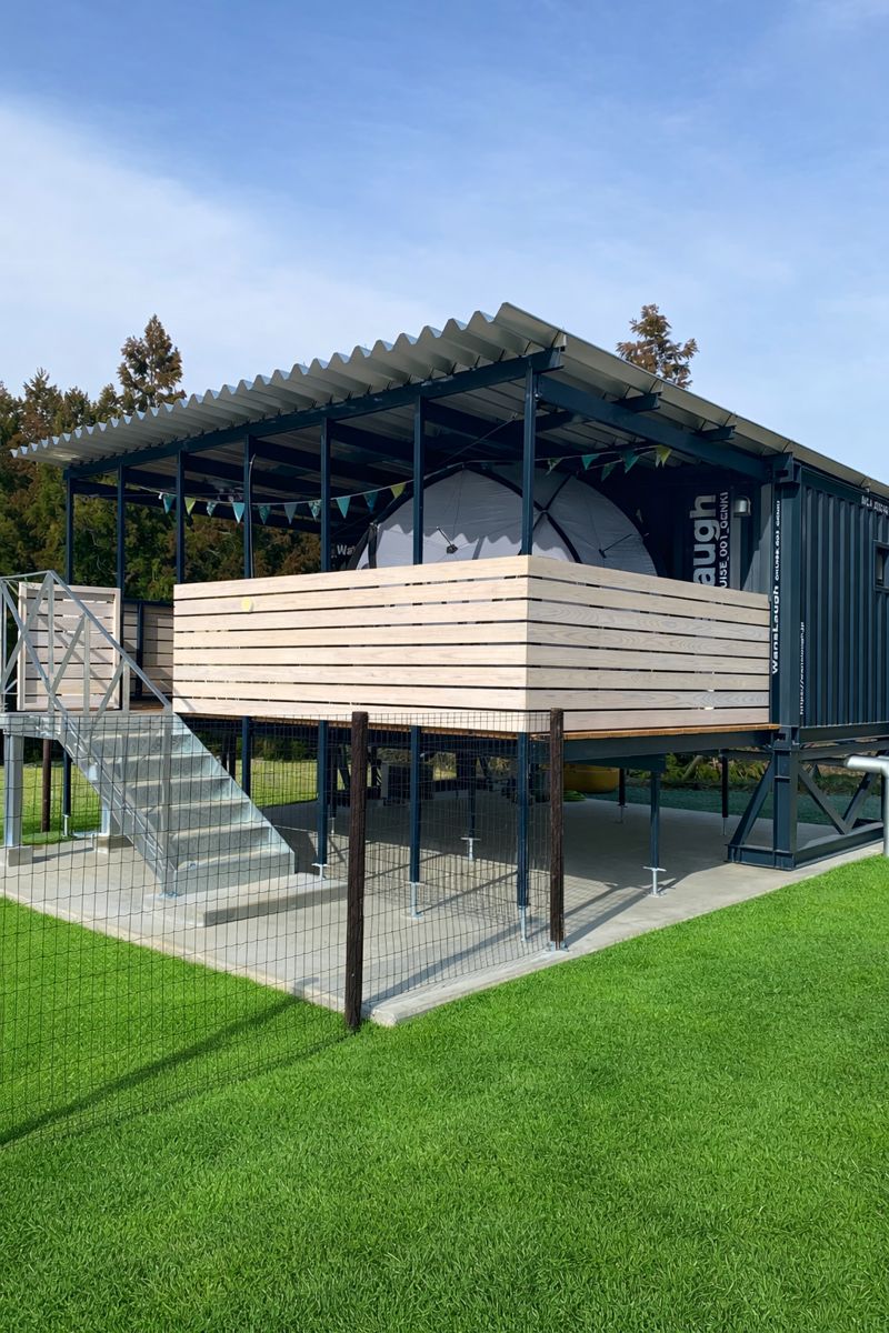 Blue shipping-container shelter on stilts with a wooden deck, metal stairs, and a fenced grassy area; a white tent is inside the deck enclosure.