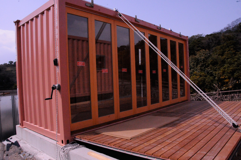 hahajima_004 - あなたがデザインする新しい建築のかたち Orange shipping-container building with full-height glass doors, mounted on a wooden deck and secured with guy wires in an outdoor setting.