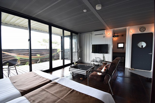 A modern hotel suite with two beds, a leather chair setup, a coffee table, and a wall-mounted TV, opening to a balcony through large glass doors.